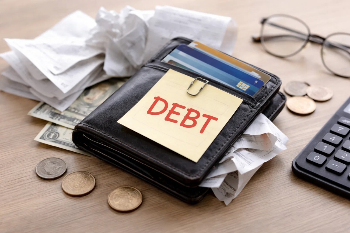 A close-up of a black wallet filled with credit cards and cash placed on a desk, surrounded by scattered receipts, coins, eyeglasses, and a calculator, with a sticky note labeled “DEBT” prominently displayed on top. This image conveys the burden of financial obligations and highlights the risks of revolving balances, aligning with credit card tips about avoiding long-term debt in the U.S. and understanding when it’s worth avoiding credit card revolving debt at all costs.